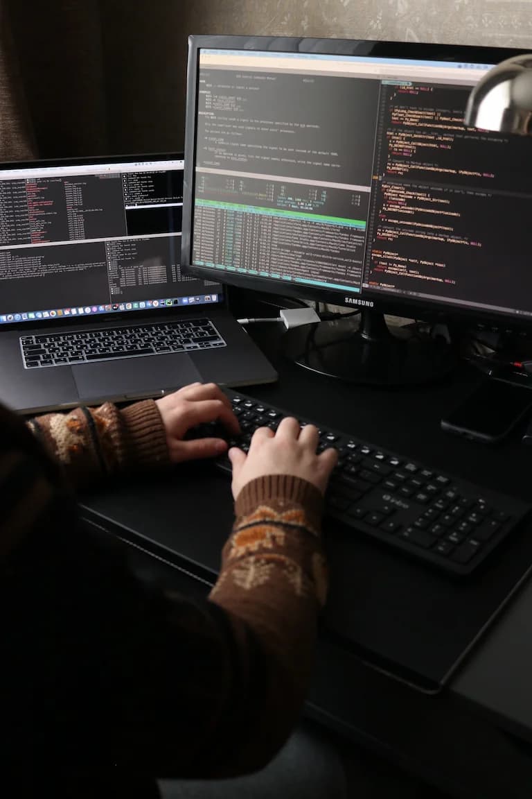 Computer screen and latop screen side by side with keyboard in front. Users hands are visible and code is on both screens